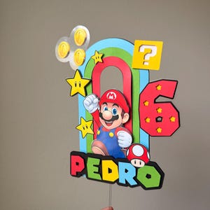 May include: A multi-layered cake topper featuring a central Mario character, a red number 6, and the name PEDRO in colorful block letters. The design includes a rainbow arch, yellow question block, gold coins, stars, and a red mushroom icon.