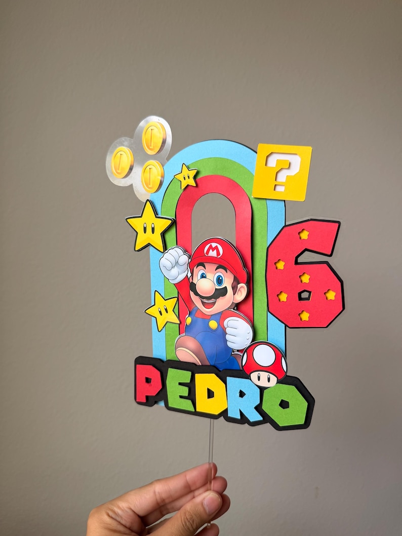 May include: A multi-layered cake topper featuring a central Mario character, a red number 6, and the name PEDRO in colorful block letters. The design includes a rainbow arch, yellow question block, gold coins, stars, and a red mushroom icon.