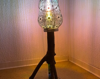 May include: A white metal lamp with a decorative pattern and a wooden base. The lamp is lit and casting a warm glow.