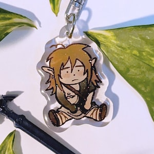 Peut inclure: Un porte-clés en acrylique transparent avec une illustration de dessin animé d'un personnage aux cheveux bruns et aux yeux verts, portant une tunique brune et tenant une épée. Le porte-clés est attaché à un anneau en métal argenté.