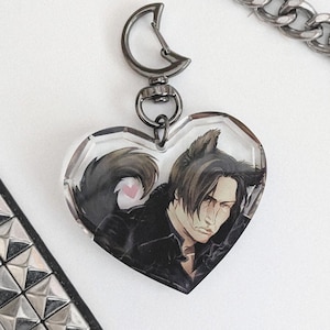 Peut inclure: Un porte-clés en acrylique en forme de cœur illustrant un personnage aux cheveux sombres avec des oreilles de loup, vêtu d'une veste noire. Le charme est fixé à un fermoir en métal sombre en forme de lune près d'une chaîne argentée et d'une sangle cloutée.