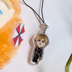 Peut inclure: Un charm en acrylique transparent représentant un personnage de dessin animé aux cheveux blonds, vêtu d'une veste et d'un pantalon noirs. Le charm est attaché à un cordon noir. Une peluche orange avec un logo rouge et blanc est en arrière-plan.