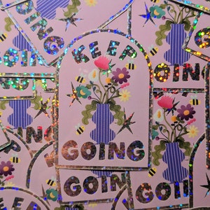 Peut inclure: Un autocollant rose avec une finition holographique présente un vase avec des fleurs et le texte "KEEP GOING". L'autocollant a une forme d'arche arrondie et une bordure argentée.