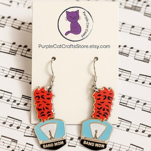 May include: A pair of dangle earrings featuring a red and black design above a light blue band hat with the words "BAND MOM" in black. The earrings are suspended from silver-colored hooks. The background includes sheet music and a logo with a purple cat.