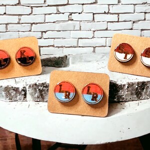 May include: Three pairs of wooden stud earrings displayed on cardboard. Each earring features a circular design with a letter or letters, and a color scheme of red, black, white, and blue. The earrings are on a white table.