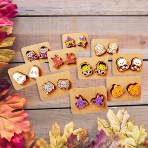 May include: A collection of Halloween-themed stud earrings on brown cardboard backing. The earrings feature various designs, including ghosts, spiders, pumpkins, bats, mummies, and Frankenstein's monster. The earrings are colorful and have a cartoonish style.