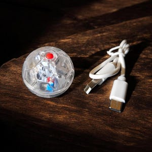 May include: A clear, soccer ball-shaped LED light with a red button on top, next to a white USB charging cable. The light is on a dark brown wooden surface.
