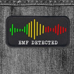 May include: Black rectangular patch with the text "EMF DETECTED" in white. The patch features a graphic of a sound wave, with green, yellow, and red vertical lines. The patch is set against a denim background.