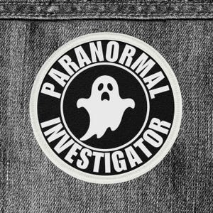 May include: A circular patch with the words "PARANORMAL INVESTIGATOR" in white, encircling a white ghost illustration on a black background. The patch is set against a gray denim fabric.
