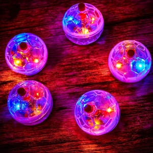 May include: Five clear, spherical LED lights with a soccer ball design. Each light emits a vibrant mix of red, blue, and yellow light. The lights are arranged on a dark wooden surface, creating a colorful display.
