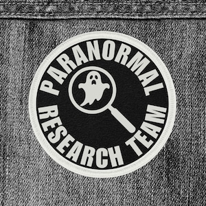 May include: A round, black and white patch with the text "PARANORMAL RESEARCH TEAM". The patch features a white ghost inside a magnifying glass. The patch is on a gray denim background.