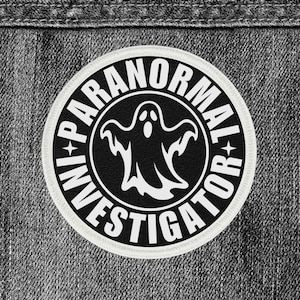 May include: A round, black and white patch with the words "INVESTIGATOR" and "PARANORMAL" encircling a white ghost graphic. The patch is set against a gray denim background.