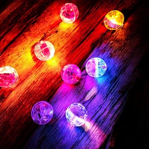 May include: A collection of glowing, spherical LED lights in various colors, including red, pink, yellow, and blue. The lights are scattered on a dark, wooden surface, creating a vibrant and festive atmosphere. These decorative lights are ideal for parties and events.