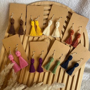Peut inclure: Assortiment de boucles d'oreilles en forme de pompon faites à la main dans différentes couleurs, notamment rose, violet, vert, bleu, jaune, marron, orange et blanc. Les boucles d'oreilles sont présentées sur du carton brun.