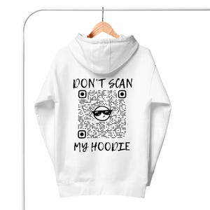 May include: White hooded sweatshirt hanging on a rack. The back displays a large QR code with the text "DON'T SCAN MY HOODIE" in black. The QR code includes a smiling face wearing sunglasses.