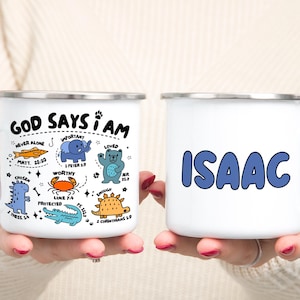 Op de afbeelding: Twee witte emaille mokken. Eén mok heeft de tekst "GOD SAYS I AM" met cartoon illustraties en bijbelverzen. De andere mok heeft de naam "ISAAC" in blauwe bubbelletters. Beide mokken hebben zilveren randen en witte handvatten.