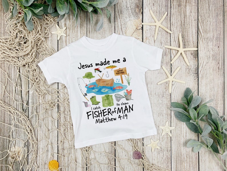 May include: White t-shirt with a colorful fishing-themed graphic. The design includes a boat, fish, fishing gear, and the text "Jesus made me a FISHER of MAN Matthew 4:19". The shirt is decorated with a fishing scene.