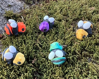 May include: Five handmade polymer clay bumblebees in various colors, including orange, purple, yellow, green, and black. The bees are sitting on a bed of green moss.