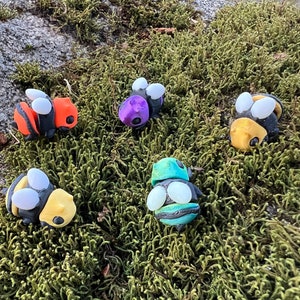 Op de afbeelding: Vijf handgemaakte bijen van polymer clay in verschillende kleuren, waaronder oranje, paars, geel, groen en zwart. De bijen zitten op een bed van groen mos.