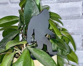 May include: A black metal silhouette of a Bigfoot standing in a pot of green leafy plants. The plant is in a cream-colored pot and is sitting on a wooden shelf.