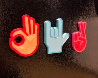 May include: Three hand gesture magnets on a black refrigerator. The magnets are made of 3D printed plastic and are in the shapes of the OK sign, the rock and roll sign, and the peace sign. The OK sign magnet is orange, the rock and roll sign magnet is blue, and the peace sign magnet is pink.