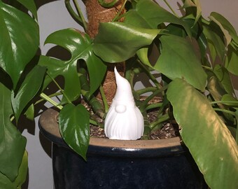 May include: A white ceramic gnome figurine with a rounded hat and a beard. The gnome is sitting in a pot of a green leafy plant.