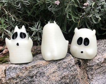 May include: Three white ghost figurines with black eyes and mouths. The ghosts are sitting on a gray rock surface.