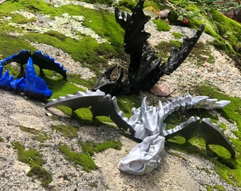 May include: Three 3D printed dragon skeletons in different poses. The skeletons are black, silver, and blue. The skeletons are on a mossy rock surface.
