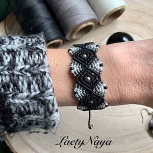 Puede incluir: Pulsera de macramé negra y gris con cuentas plateadas. La pulsera está atada con un cordón negro y tiene una cuenta plateada en el extremo.