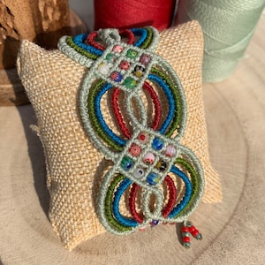 Könnte beinhalten: Ein farbenfrohes Makramee-Armband mit grünen, blauen und roten Schnüren und Glasperlen. Das Armband wird auf einer beigefarbenen Stoffoberfläche präsentiert.