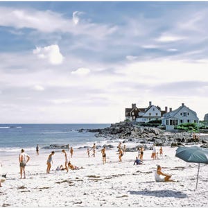 May include: A vintage photograph captures a sunny beach scene with people enjoying the day. The sky is blue with scattered clouds. Houses are visible in the background. Beach umbrellas provide shade on the sandy shore.