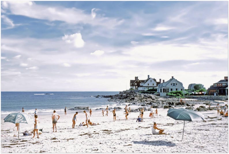 May include: A vintage photograph captures a sunny beach scene with people enjoying the day. The sky is blue with scattered clouds. Houses are visible in the background. Beach umbrellas provide shade on the sandy shore.