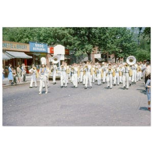Puede incluir: Una banda de música con uniformes blancos con detalles dorados e instrumentos desfila por una calle. Los miembros de la banda tocan instrumentos de viento metal. Los espectadores se alinean en las aceras. Una panadería y una zapatería son visibles en el fondo.