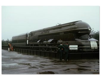 Puede incluir: Una locomotora de vapor negra con un diseño aerodinámico, de pie en una plataforma con una valla de metal negro. Dos personas están de pie frente a la locomotora, mirándola.