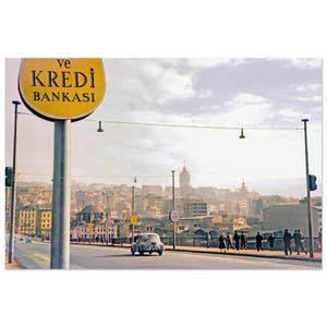 May include: Vintage photograph of a city street scene with a yellow sign that reads "ve KREDI BANKASI". A classic car drives on the road, and people walk on the sidewalk. The background features buildings and a cloudy sky.