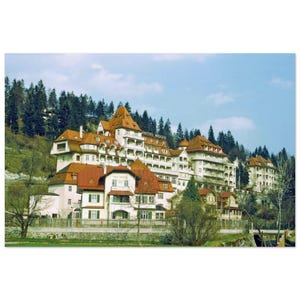 Puede incluir: Fotografía vintage de un gran hotel en una ladera. El edificio tiene paredes blancas, tejados de tejas rojas y muchas ventanas. El hotel está rodeado de árboles verdes y un cielo azul. El nombre del hotel es Hotel Wigger.