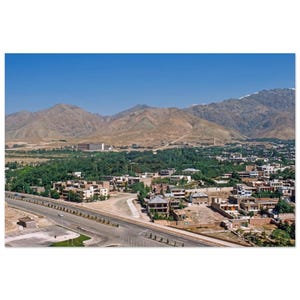 May include: A wide-angle view of a town nestled at the base of large, arid, tan-colored mountains under a clear blue sky. A multi-lane paved road runs through the foreground, with residential buildings and dense green trees filling the valley.