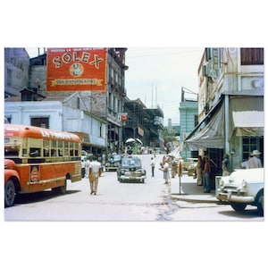 May include: A vintage street scene featuring a red bus, classic cars, and pedestrians. A large sign for "SOLEX" paint is displayed above the street. Buildings line the road, and people are walking on the pavement.
