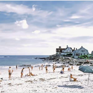 May include: A vintage photograph of a beach scene with people enjoying a sunny day. The sky is blue with scattered clouds. Several people are on the sandy beach, some under umbrellas. Houses are visible in the background.