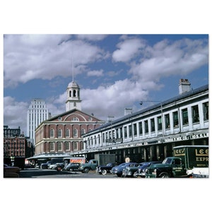 Pode incluir: Uma fotografia antiga do Faneuil Hall Marketplace em Boston, Massachusetts. O edifício de tijolos com uma torre de relógio está no centro da imagem. Há vários carros estacionados em frente ao edifício, incluindo um caminhão de entrega verde com as palavras "Lee's Express" no lado.