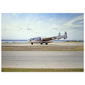 May include: A vintage silver airplane, marked "UNITED STATES MARINES", is on a runway. The aircraft has a star emblem and the number "MZ" on its tail. The sky is blue with scattered clouds, and the landscape includes a body of water and grassy areas.