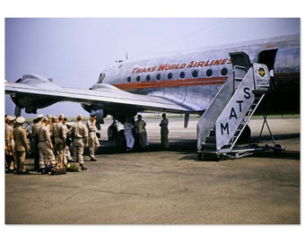 Puede incluir: Un grupo de personas con uniformes militares se encuentran en la pista de aterrizaje frente a un avión plateado de Trans World Airlines. El avión tiene una franja roja y las palabras "Trans World Airlines" impresas en el lateral. Una escalera de metal está unida a la puerta del avión.
