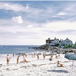 May include: A vintage photograph of a beach scene with people enjoying the sun. The image shows a sandy beach, blue ocean, and a cloudy sky. Several people are on the beach, some under umbrellas, and houses are visible in the background.