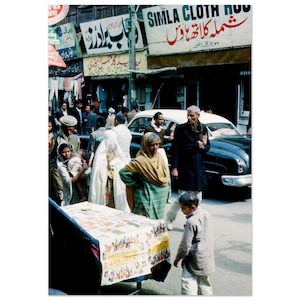 May include: A busy street scene in India with people walking and shopping. A vintage car is parked on the street. A sign above a shop reads "SIMLA CLOTH HOUSE".