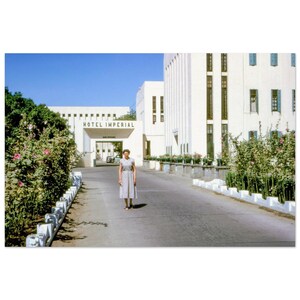 May include: A woman stands on a paved driveway leading to the Hotel Imperial. The hotel is a white building with a sign that reads "HOTEL IMPERIAL". Green bushes with pink flowers line the driveway. The sky is blue.