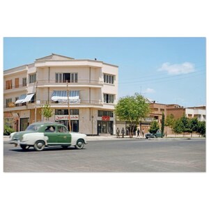 May include: A vintage photograph of a street scene featuring a two-tone green and white classic car driving on a paved road. A multi-story building with awnings and storefronts is in the background under a clear blue sky.