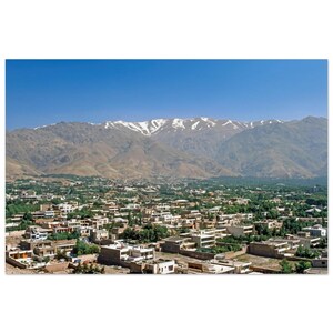 May include: A wide-angle landscape view of a sprawling city nestled at the base of a massive mountain range. The mountains feature rugged, tan-colored slopes with patches of white snow near the peaks under a clear, bright blue sky.
