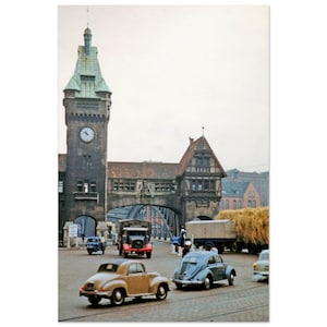 May include: A vintage photograph depicting a European city scene with a clock tower, arched bridge, and classic cars. The image has a muted colour palette, focusing on architectural details and vehicles from a past era.