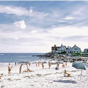 May include: A vintage photograph captures a sunny beach scene with people enjoying the day. The sky is a mix of blue and white clouds. Several houses sit on a rocky outcrop. Beach umbrellas provide shade as people play and relax on the sand.