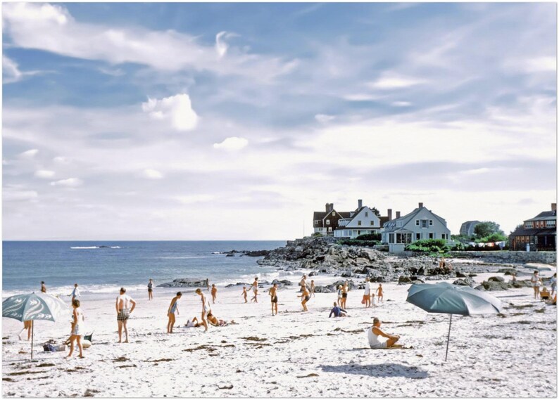 May include: A vintage photograph captures a sunny beach scene with people enjoying the day. The sky is a mix of blue and white clouds. Several houses sit on a rocky outcrop. Beach umbrellas provide shade as people play and relax on the sand.